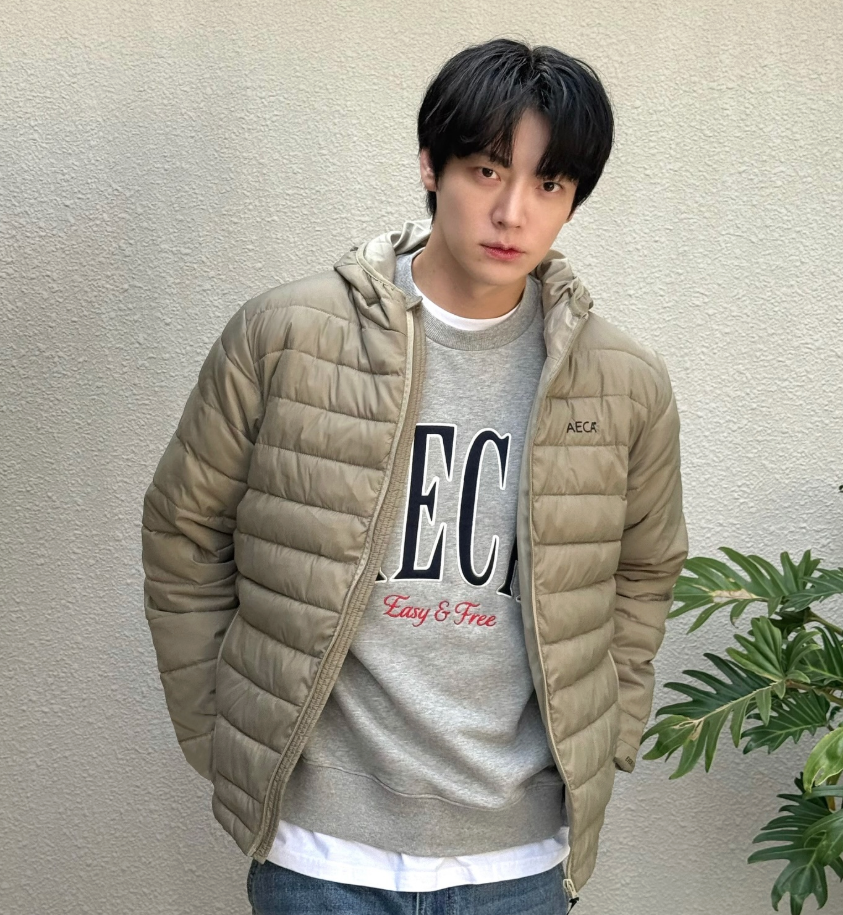회색 맨투맨 티셔츠 위에 베이지색 AECA 패딩 점퍼를 입고 주머니에 손을 넣은 채 카메라를 정면으로 응시하고 있는 배우 안재현의 모습