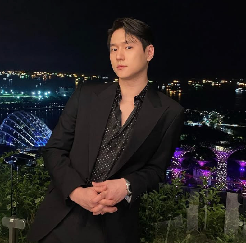 화려한 도시의 야경과 바다를 배경으로 검은색 수트와 도트 무늬 셔츠를 입고 난간에 기대어 정면을 응시하는 배우 고경표의 모습