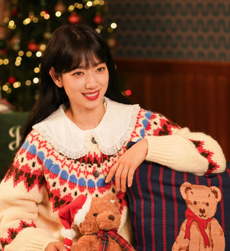 크리스마스트리와 따뜻한 조명이 가득한 배경에서 레이스 깃이 달린 노르딕 패턴 니트를 입고 곰 인형 쿠션 곁에서 밝게 웃고 있는 배우 박신혜의 모습
